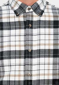 Checkered marškiniai, kuriuose derinami juoda, balta ir rusva spalvos. Marškiniai su nusileidžiančiu apykaklės dizainu, rudos spalvos sagomis ant tekstūruoto medvilninio audinio.