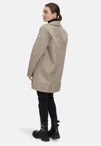 Beige vattentät kappa med krage och avslappnad passform, med sidofickor. Bärs över svarta byxor och kraftiga svarta stövlar.