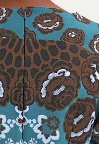 Flerfärgad tyg med bruna och teal blommönster samt leopardmönster. Har en bakre dragkedja, strukturerad yta och kontrasterande blå accenter.