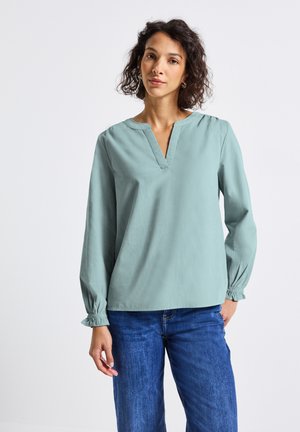 Vrouw met krullend haar die een lichtblauwe blouse met lange mouwen en een blauwe spijkerbroek draagt, staande tegen een effen witte achtergrond.