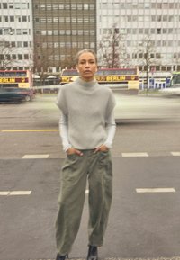 Person mit grauem ärmellosem Pullover und grüner Hose steht mit den Händen in den Taschen auf der Stadtstraße, im Hintergrund Busse und Gebäude.