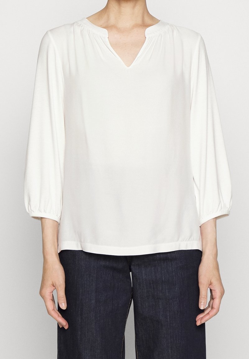 Camicia bianca con scollo a V, maniche 3/4 e dettagli arricciati sulle spalle. Tessuto liscio, vestibilità comoda, abbinata a pantaloni di jeans scuri.