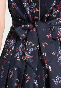 Marinblå blommig klänning med knytmidja, som har färgglada blommor och fjärilar. Slätt tyg med dragkedja bak.