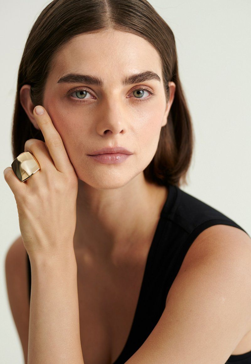 Femme aux cheveux courts bruns et aux yeux verts portant un haut noir sans manches, reposant son visage sur sa main avec une grosse bague en or à son doigt.