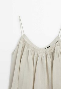 Robe sans manches beige clair avec fines bretelles et tissu froncé le long du décolleté, présentée sur un fond blanc.