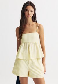 Gelb gestreiftes Peplum-Top mit dünnen Trägern, kombiniert mit passenden Shorts. Der Stoff ist leicht und hat strukturierte, vertikale Streifen.
