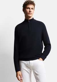 Marineblauer Strickpullover mit einem Viertelreißverschluss und strukturiertem Muster. Lange Ärmel mit gerippten Bündchen und Saum, kombiniert mit weißen Hosen.