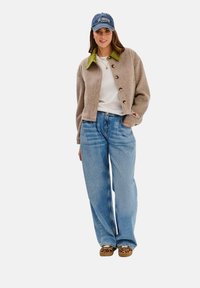 Beżowy krótki kardigan z drewnianymi guzikami, zielonym kołnierzykiem; biała koszula pod spodem; jasnoniebieskie luźne dżinsy; brązowe buty w panterkę; niebieska czapka.