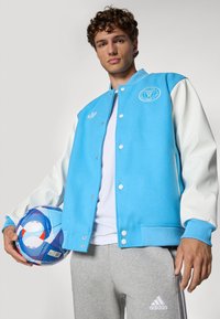 Light blue varsity jacket with white sleeves, featuring a logo on the chest and ribbed cuffs. Gray sweatpants and a colorful soccer ball.