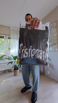 Packaging negro con "Visionary" en texto blanco y en negrita. La persona que lo sostiene lleva jeans azules y zapatos negros. Ambiente interior con plantas.