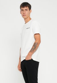 Hvid bomulds t-shirt med korte ærmer, prydet med et lille sort "REPLAY" logo på brystet. Sammen med sorte bukser.