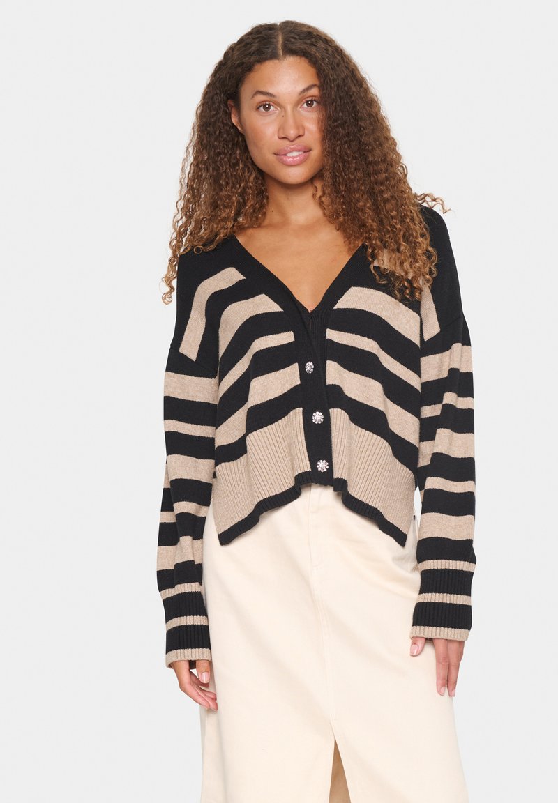 Cardigan rayé noir et beige avec un col en V, un ourlet côtelé et une fermeture à trois boutons ornée de fleurs. Porté par-dessus une jupe légère.