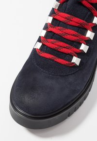 Námořnické semišové tenisky s červenými a černými vzorovanými tkaničkami, stříbrnými obdélníkovými očka a silnou černou gumovou podrážkou.