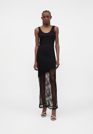 Femme portant une robe noire sans manches avec un ourlet asymétrique et une jupe en dentelle transparente, associée à des talons blancs à brides, debout devant un fond uni.