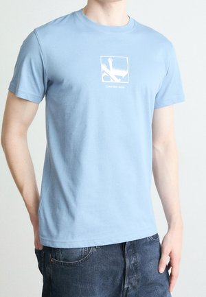 Homme portant un t-shirt Calvin Klein Jeans bleu clair avec un motif blanc et un jean bleu foncé sur un fond uni.