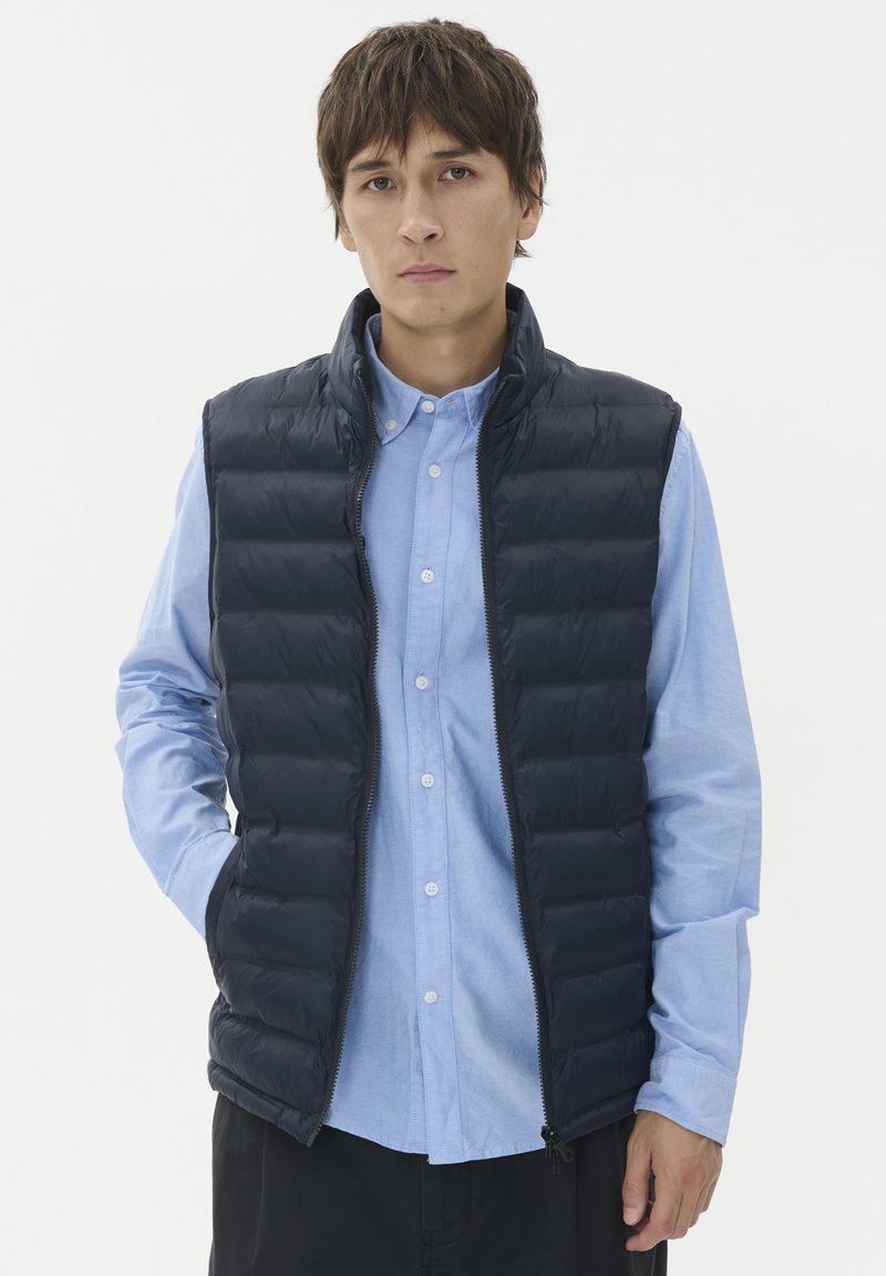 Pufferväst i mörk marinblå, med kviltade sektioner, ståkrage och dragkedja fram. Buren över en ljusblå skjorta med knappar.