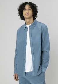 Chemise boutonnée en velours côtelé bleu clair avec une coupe décontractée, manches longues et col classique, portée sur un t-shirt blanc.
