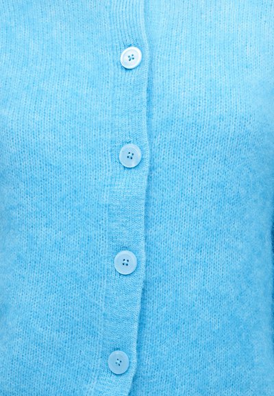 Κοντινό πλάνο ενός ελαφρώς γαλάζιου πλεκτού ζακέτας με τέσσερα διαφανή μπλε κουμπιά δεμένα σε κάθετη σειρά.