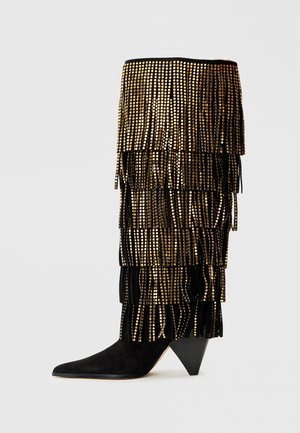 Botte en suede noire avec des franges dorées, dotée d'un bout pointu et d'un talon triangulaire. Des disques brillants embellissent la partie supérieure.