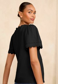 Femme avec les cheveux en bun portant un haut noir à manches courtes avec des bords festonnés, des boucles d'oreilles en or, regardant par-dessus son épaule sur un fond beige.