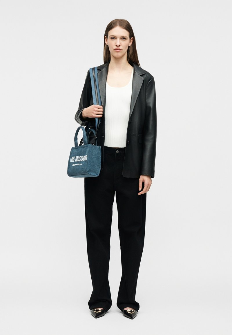 Γυναίκα ντυμένη με μαύρη δερμάτινη ζακέτα, λευκή μπλούζα, μαύρο παντελόνι, μαύρα μυτερά παπούτσια με χρυσές λεπτομέρειες, κρατώντας μπλε τζιν τσάντα Love Moschino.