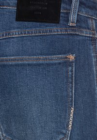 Bakfickan på blå denimjeans med beige sömmar och en svart läderlapp med texten "Stockholm Melbourne 2009."