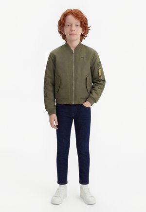 Garçon aux cheveux roux portant une veste bomber vert olive, un jean bleu foncé et des baskets blanches, debout devant un fond blanc.