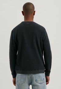 Uomo in piedi rivolto di spalle, con indosso un maglione scuro a maniche lunghe e jeans in denim azzurro chiaro, su uno sfondo uniforme.