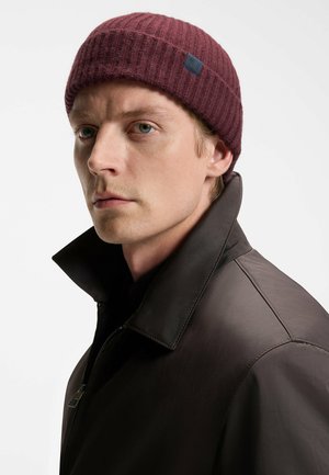 Burgundy ribbad mössa med en liten mörkblå etikett; bärs med en brun läderjacka med hög krage och dragkedjestängning.