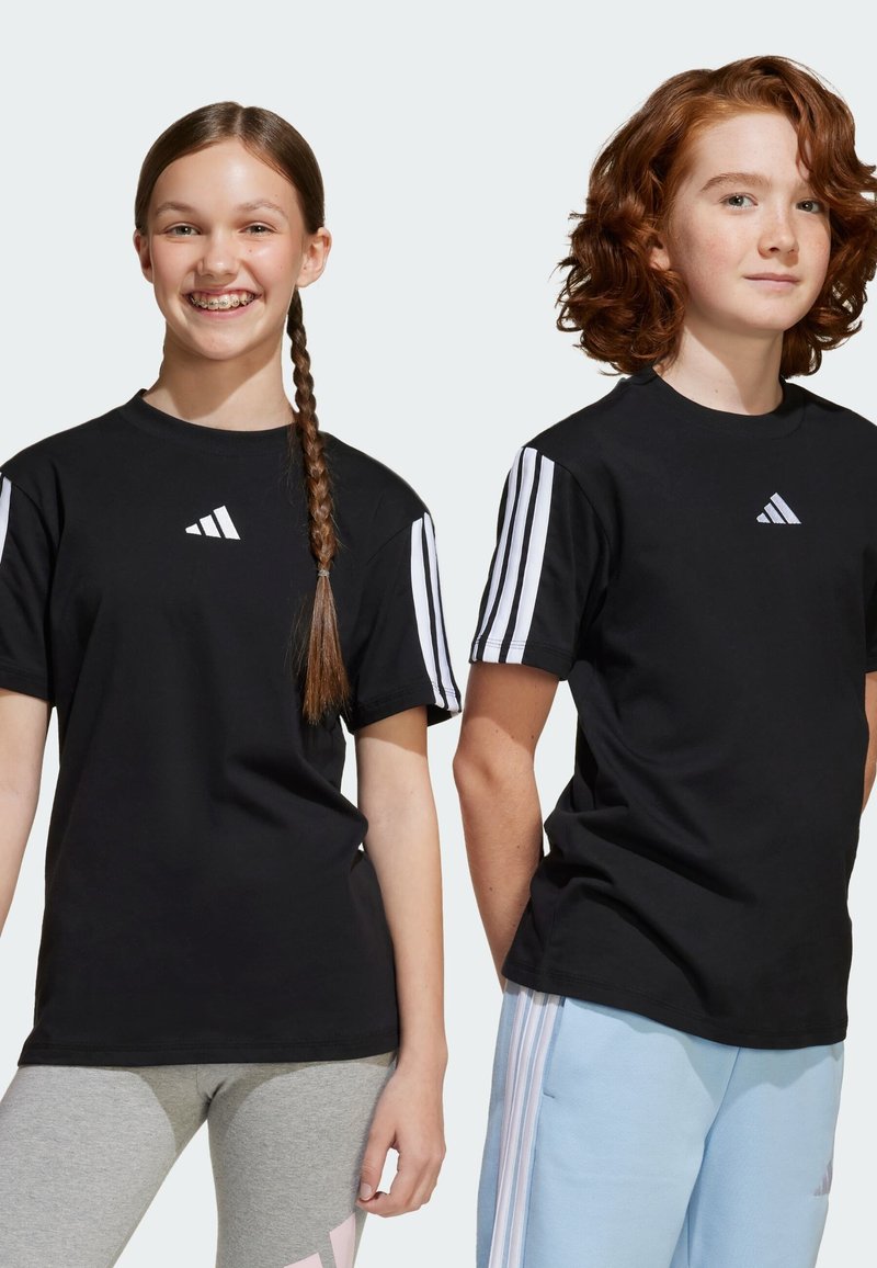 T-shirts noirs avec des détails à trois rayures blanches sur les manches et un logo sur la poitrine. Deux modèles jeunes se tiennent côte à côte, l'un en leggings gris, l'autre en pantalon bleu clair.