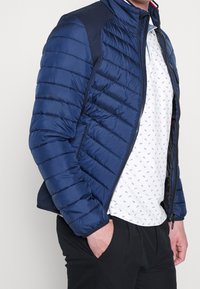 Mann trägt eine blaue gesteppte Jacke mit Reißverschluss über einem weißen gemusterten Hemd und schwarzen Hosen, mit einer Hand in der Tasche vor einem schlichten Hintergrund.