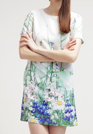 Kobieta w krótkiej białej sukience z kolorowym kwiatowym wzorem, z założonymi skrzyżowanymi ramionami, stojąca na tle jasnego, jednolitego tła.