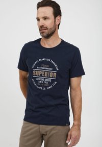 Marineblå bomulds T-shirt med rund hals og korte ærmer, prydet med et trykt logo i beige og hvidt med cirkel accenter på fronten.