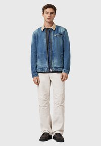 Denim-takki, jossa on liukuvärjätty sininen pinta, beigenvärinen kaulus, vetoketjusuljenta ja sivutaskut. Yhdistetty vaalean kermaisiin corduroy-housuihin ja tummiin kenkiin.