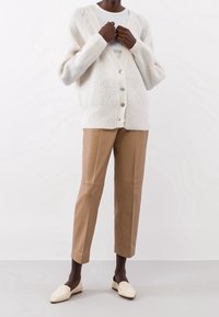 Cremfarbener Cardigan mit V-Ausschnitt, Knopfleiste und geripptem Muster, kombiniert mit hellbraunen Lederhosen und weißen Slippern.