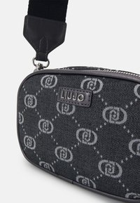 Bolso de tela negro con un patrón geométrico gris, cierre con cremallera y un asa de cuero negro con un detalle del logo.