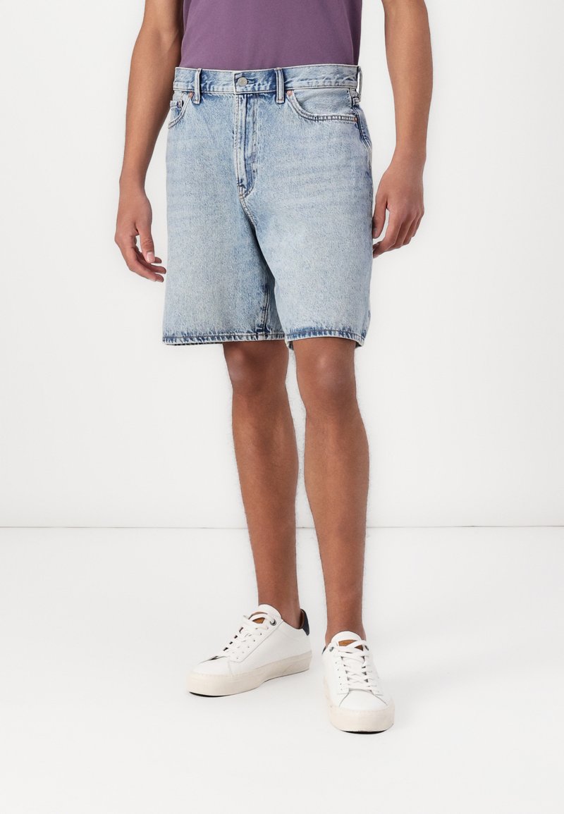 Shorts de mezclilla en azul claro, con un corte relajado, dobladillo deshilachado y diseño de cinco bolsillos, combinados con deportivas blancas y una camiseta morada.