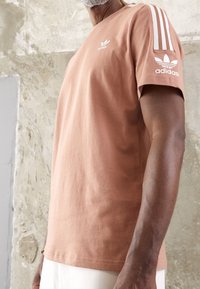 Persikofärgad bomullströja med vit Adidas-logotyp på bröstet och tre ränder på axeln. Kort ärm och avslappnad passform.