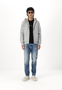 Grå zip-hoodie med lommer, beige foring, sort t-shirt, blå jeans, cremede sneakers og runde solbriller. Ensfarvet baggrund.