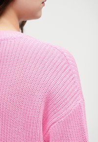 Roza pletena jopica z teksturiranim vzorcem. Ima sproščen kroj in okroglo ovratnico, ki razkriva področje zadnjega ramena.