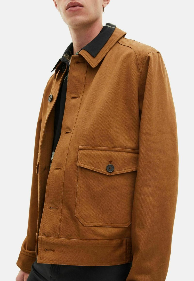 Veste en toile marron avec un col noir, fermetures à boutons et une poche avant. Elle présente une texture lisse et un design structuré.