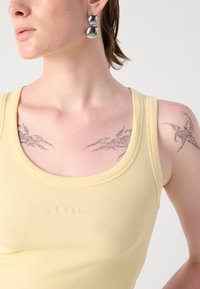 Ribbed, sleeveless yellow tank top with a round neckline and BOSS logo. Model wears large silver dangle earrings and displays tattoos on shoulders.