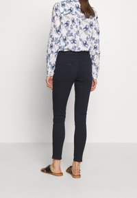 Mujer de pie contra un fondo blanco, vistiendo pantalones skinny negros, blusa blanca con estampado floral azul y sandalias negras de punta abierta.