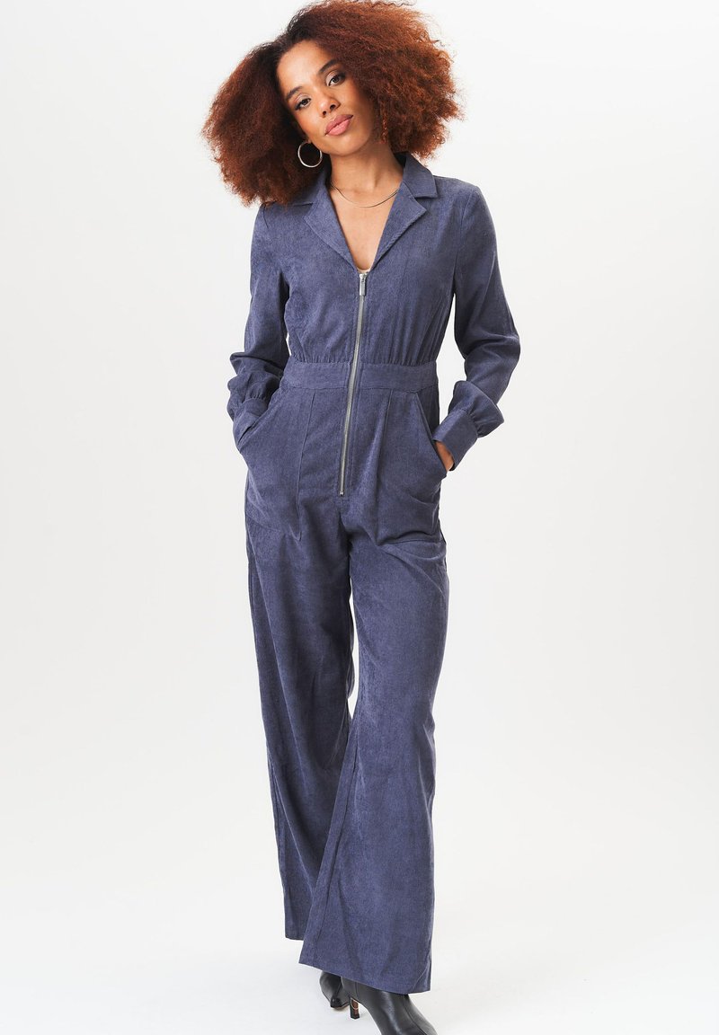 Navy blue corduroy jumpsuit with a zip front, collar, long sleeves, side pockets, and wide-leg design. Smooth, textured fabric.