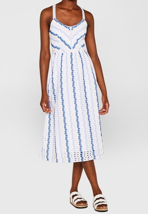 Kobieta ubrana w białą, bezrękawnikową sukienkę midi z niebieskimi pionowymi paskami i okrągłymi wzorami, w parze z białymi sandałami na podwójne paski.