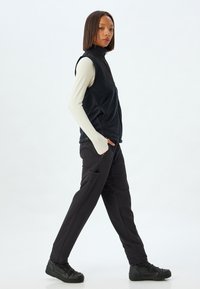 Sort fleece vest med høj krave, frontlynlås og sidelommer, båret over en cremet langærmet top og parret med sorte bukser og sorte sko.