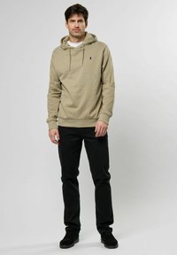 Beige katoenen hoodie met trekkoord, geribde manchetten en een klein logo. Gecombineerd met zwarte jeans en zwarte schoenen. Eenvoudig, casual ontwerp.