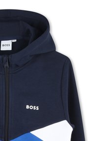 Sweat-shirt à capuche bleu marine avec des accents géométriques blancs et bleus ; possède une fermeture éclair à l'avant et une étiquette logo sur la poitrine.