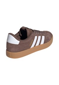 Scarpa sportiva in suede marrone con tre strisce bianche, suola in gomma naturale e dettagli beige. Presenta chiusura con lacci e colletto imbottito.