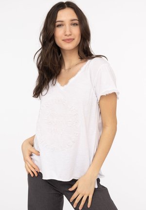 Weißes Baumwoll-T-Shirt mit V-Ausschnitt, Fransenärmel und strukturiertem Schriftzug auf der Vorderseite, kombiniert mit dunkelgrauen Hosen.